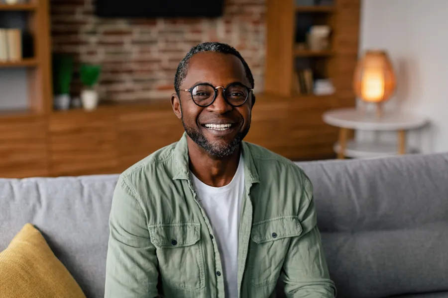 Andropause-Doctor A middle aged black man in a green button up shirt sits on his couch smiling, happy with his treatment for andropause and low testosterone from Optimal Wellness Medical Group in Grand Rapids.