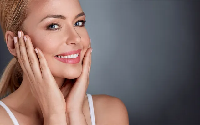 Facial-Clinic A middle-aged blonde woman with beautiful skin smiles and holds her face. Get facial treatments from Optimal Wellness Medical Group in Grand Rapids.