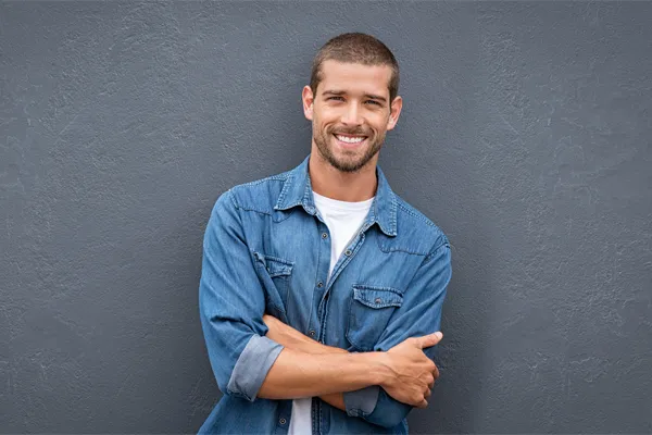 Hormones-for-Men-Doctor A man in a denim shirt stands smiling against a gray-blue wall, pleased with his testosterone hormone therapy from Optimal Wellness Medical Group in Grand Rapids.