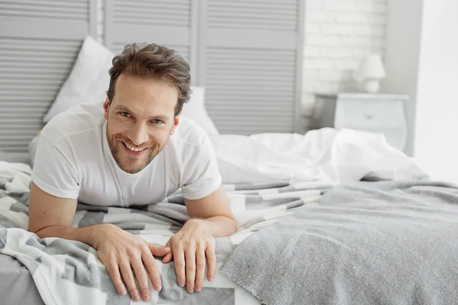 Low-Libido-in-Men-Doctor A dirty-blonde haired man lays on his stomach in bed smiling. Schedule treatment for low libido in men from Optimal Wellness Medical Group in Grand Rapids.