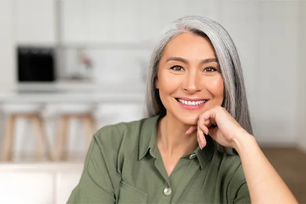 Menopause-Clinic An asian woman with long gray hair and a green shirt sitting and smiling on her couch, happy with her menopause care from [NAME} in Grand Rapids.