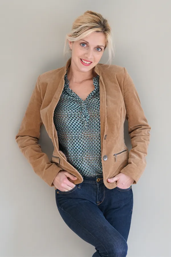 Metabolic-Syndrome-Treatment A smiling blonde woman in a tan leather jacket leans against a gray wall. Schedule metabolic syndrome from Optimal Wellness Medical Group in Grand Rapids.