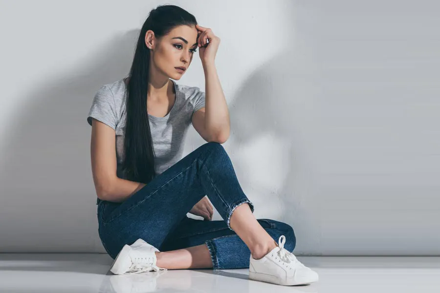 PMOS-Doctor A young brunette woman in a gray t-shirt and jeans sits against a gray wall with her head resting on her hand and a distressed expression. Get help for Polyendocrine Metabolic Ovarian Syndrome (PMOS) from Optimal Wellness Medical Group in Grand Rapids.