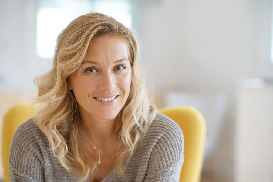 Perimenopause-Doctor A blonde woman wearing a a gray sweater sits inside in a yellow chair, smiling and happy with her perimenopause treatment from Optimal Wellness Medical Group in Grand Rapids.