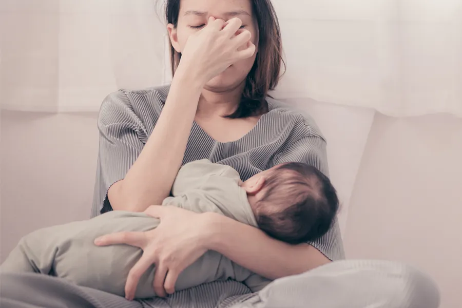 Postpartum-Hormone-Imbalance-Clinic A woman nursing an infant on couch rubs her eyes with exhaustion. Get treatment for postpartum hormone imbalance from Optimal Wellness Medical Group in Grand Rapids.