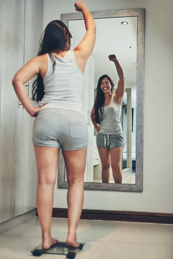 Tirzepatide-Treatment A woman in workout clothes stands on a scale looking in the mirror and pumping her hands in celebration of her successful Tirzepatide treatment from Optimal Wellness Medical Group in Grand Rapids.