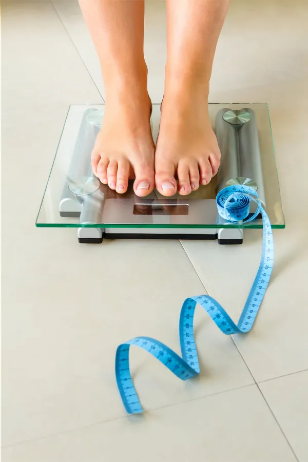 Weight-Loss-Resistance-Treatment Close-up of a woman's feet standing on a scale, with measuring tape by her toes, getting treatment for weight loss resistance from Optimal Wellness Medical Group in Grand Rapids.