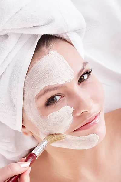 facials-grand-rapids Close up of a woman with hair wrapped in a white towel softly smiling at the camera while having a facial mask applied. Schedule a personalized facial with Optimal Wellness Medical Group in Grand Rapids.